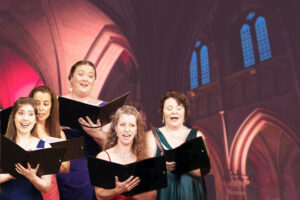 A group of singers wearing colourful evening gowns stand with their choir folders. The background is a vaulted church ceiling lit in Christmassy red tones.